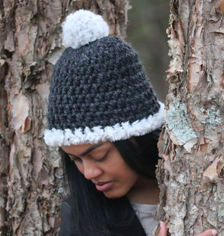 Woman wearing a charcoal satin lined winter hat with a white faux fur trim and pom.