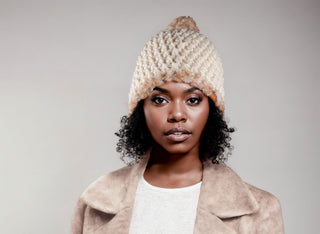 Woman wearing a cream satin lined winter hat with a brown and white faux fur pom and trim.